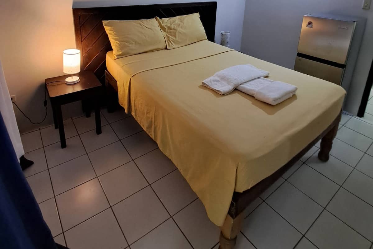 Ein ordentlich bezogenes Bett mit gelber Bettdecke und Kissen, eine Nachttischlampe und ein kleiner Tisch mit einer Tasse.