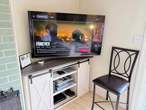 A television mounted on a wall with a wooden shelf below displaying books and a gaming console.