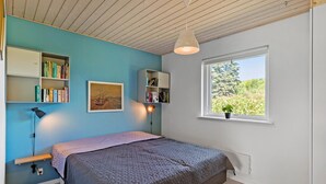 Ein Schlafzimmer mit Bett, Bücherregal, Schreibtisch, einem Fenster mit Blick auf Grünflächen und einer Pendelleuchte.