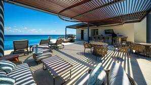 A sunlit outdoor seating area with lounge chairs and tables, overlooking a beach and ocean. - Marila - Luxury 3 BR - Ocean View & Pool (Playa del Carmen)