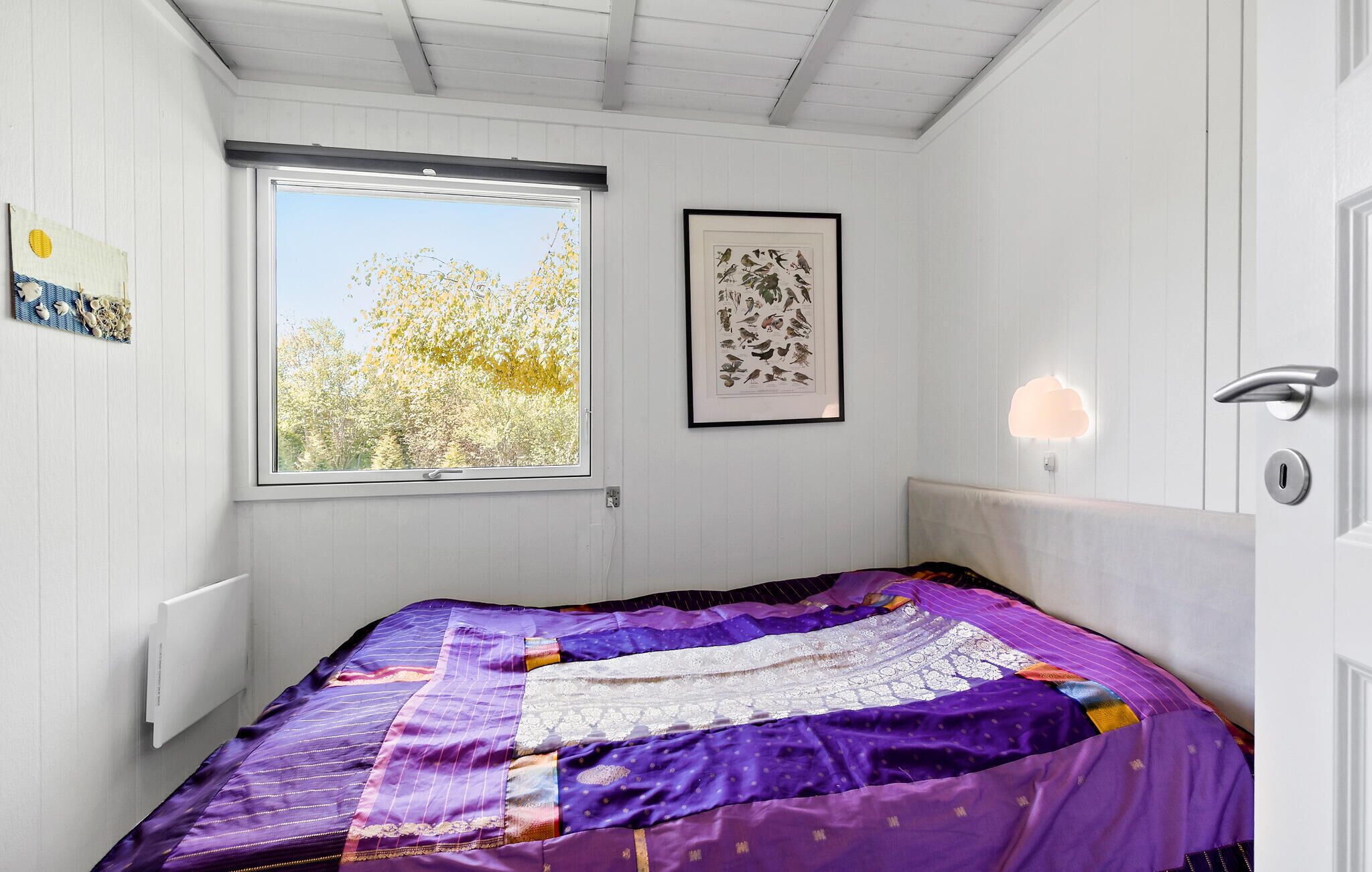 A bedroom with a large window, a framed bird illustration, and a bed with a purple quilt.