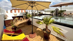 Une terrasse sur le toit avec une piscine à débordement en verre, des sièges extérieurs et des parasols.