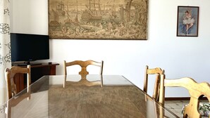 Un comedor con una mesa de vidrio, sillas de madera, un cuadro y un televisor sobre un soporte.