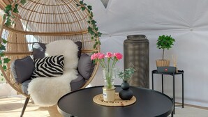 Habitació amb taula rodona negra, cadira de vimetes amb coixins, cadira penjant i un vas alçat amb flors roses.