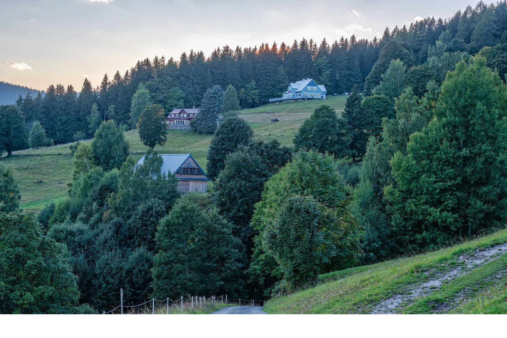 Un paysage rural avec une maison située sur une colline, entourée d’arbres denses et sous un ciel dégagé.