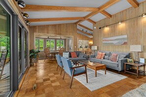 A spacious living room with a wooden floor, a grey sofa, blue armchairs, and a dining area with wooden furniture.