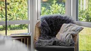 Ein Korbstuhl mit flauschiger, grauer Decke und gestreiftem Kissen, platziert vor einem Fenster mit Blick auf Grünflächen.