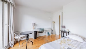 A hotel room with a bed, a desk with a chair, a small table, a mirror, and a wardrobe.