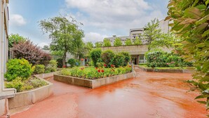 A courtyard with a paved area, surrounded by various plants and flowers, and a building in the background.