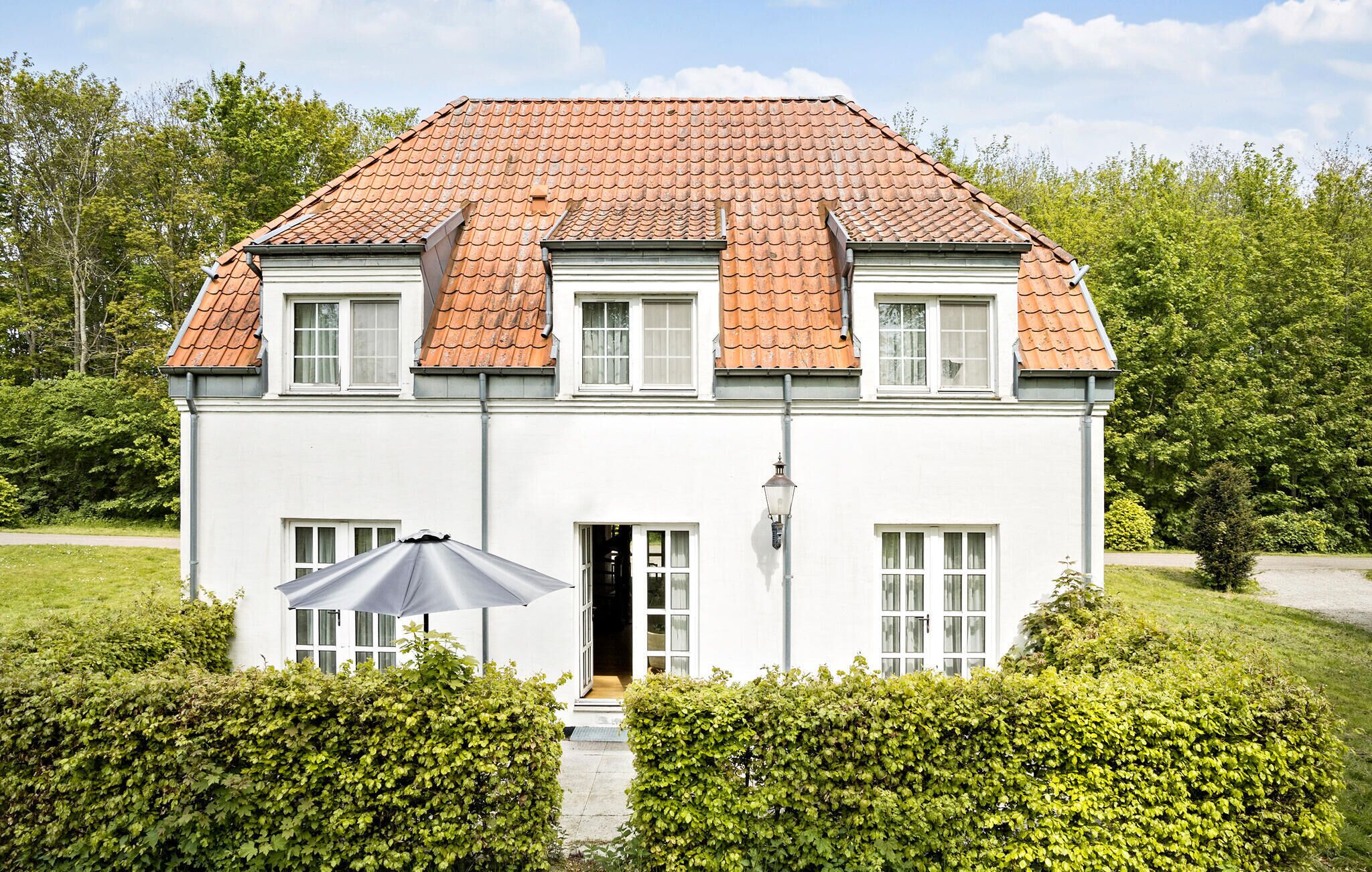 A two-story house with a red-tiled roof, white exterior walls, and a front door. There is a small garden with green hedges in front of the house.