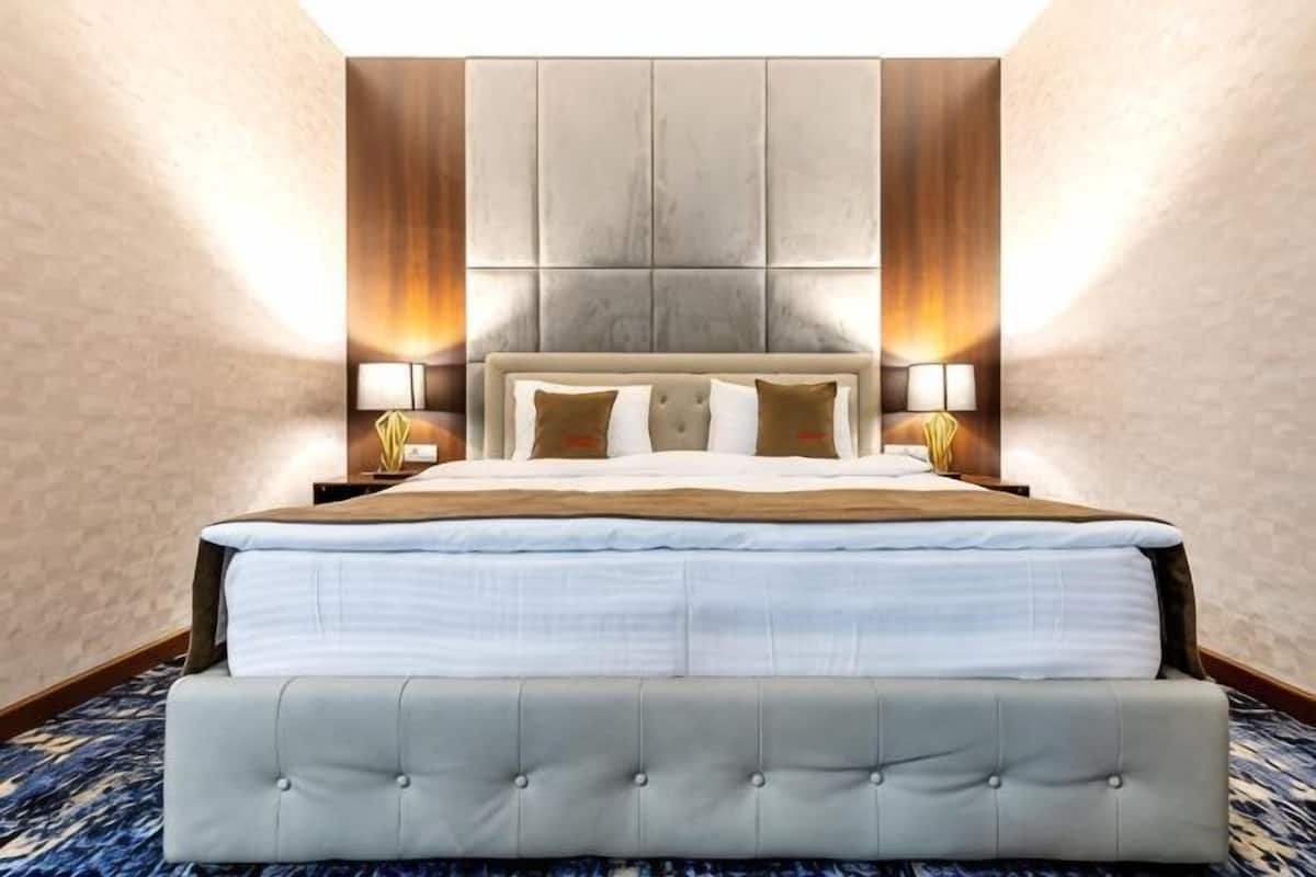 Uma cama grande com cabeceira acolchoada, duas mesas de cabeceira com abajures e um tapete azul estampado.