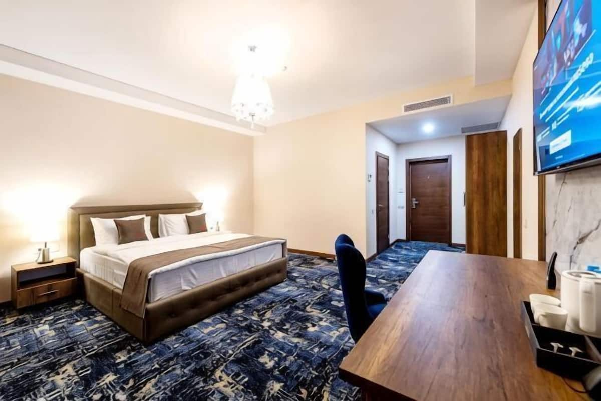 Quarto de hotel com cama, mesas de cabeceira, escrivaninha, cadeira e televisão fixada na parede.