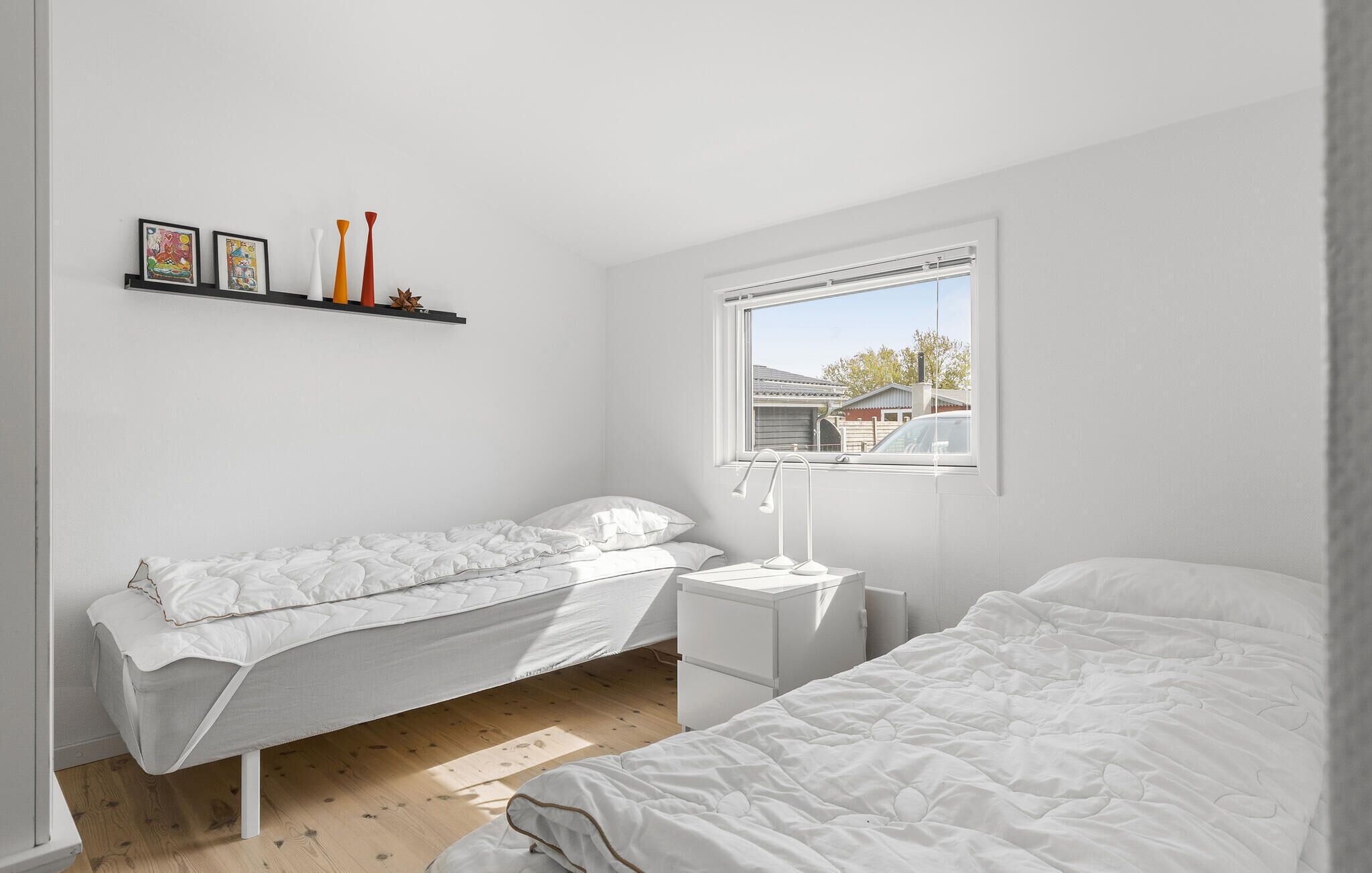 Une chambre avec deux lits, une table de chevet, une lampe et une fenêtre donnant sur une maison et une voiture à l’extérieur.