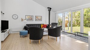 Un salon moderne avec un canapé en cuir noir, deux fauteuils gris, un plancher en bois, un meuble TV noir, une horloge et deux œuvres d’art encadrées.