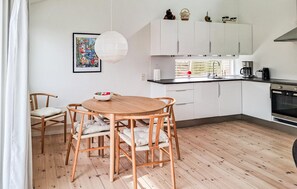 Una cocina moderna con armarios blancos, una mesa de comedor de madera con sillas, un colgante de luz y una ventana con persianas.