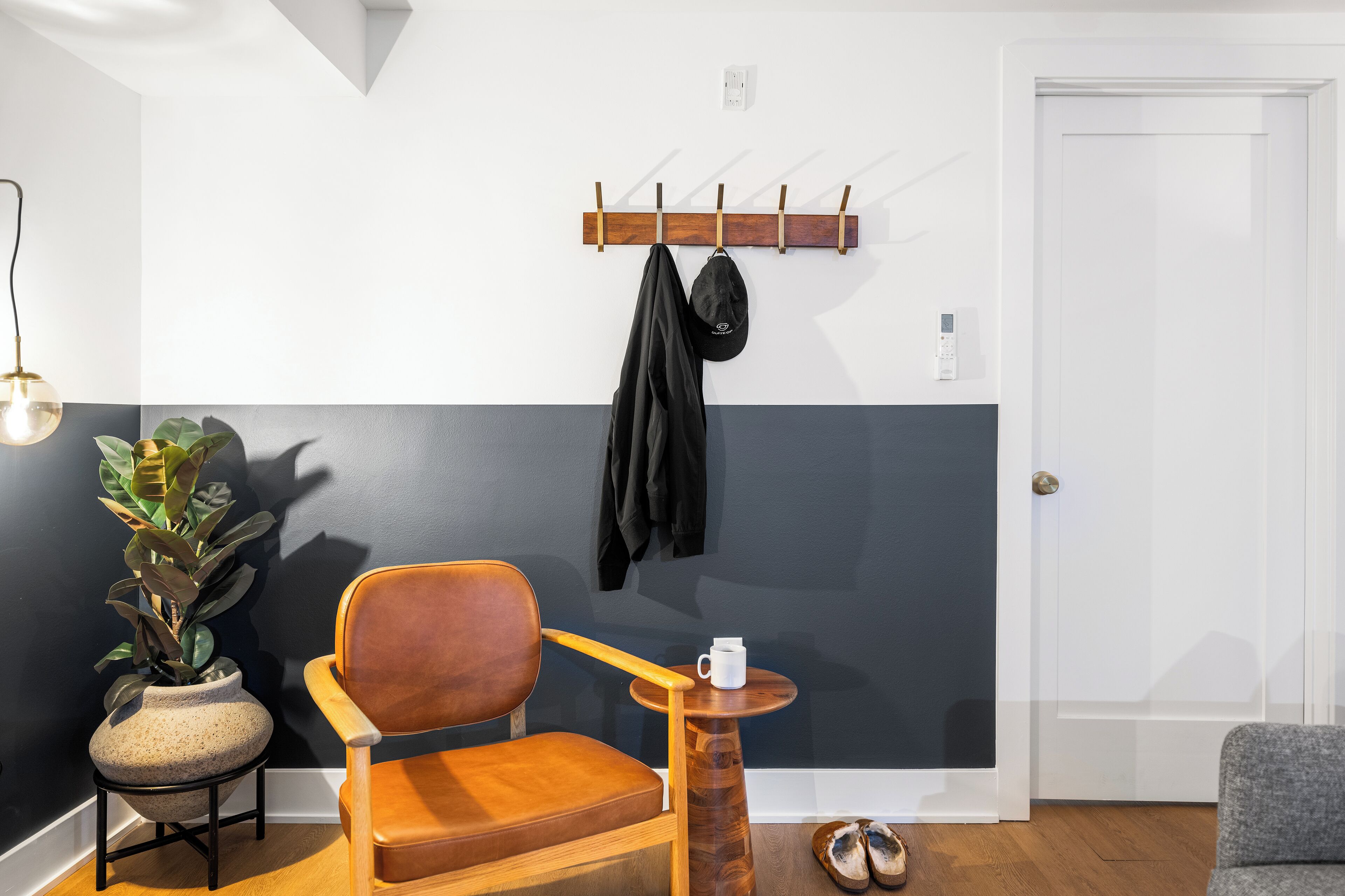 A room with a wooden chair, a small round table with a white mug, a coat rack with a jacket, and a potted plant.