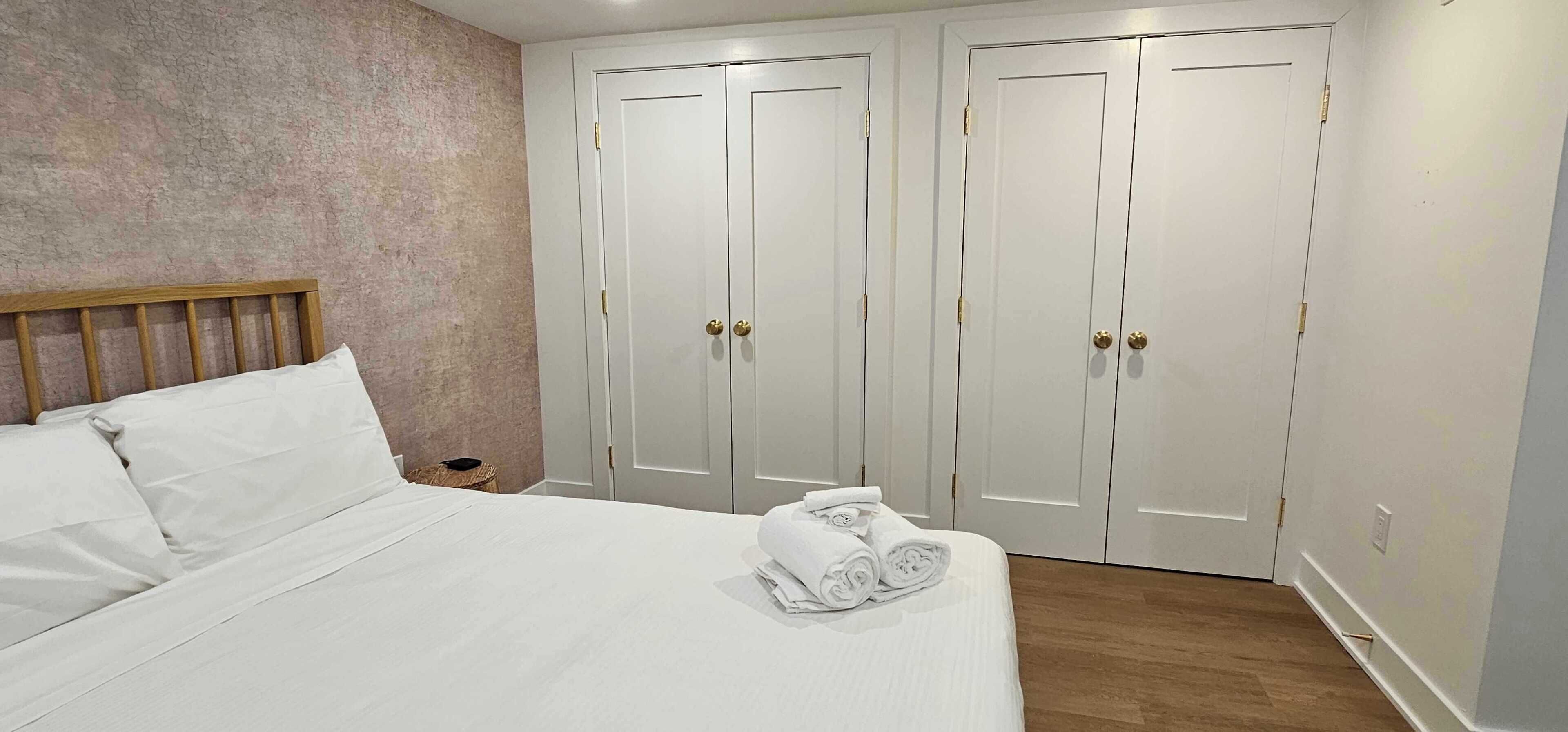 Een net opgemaakt bed met witte lakens en een opgevouwen handdoek. Op de achtergrond twee witte deuren met gouden handgrepen.