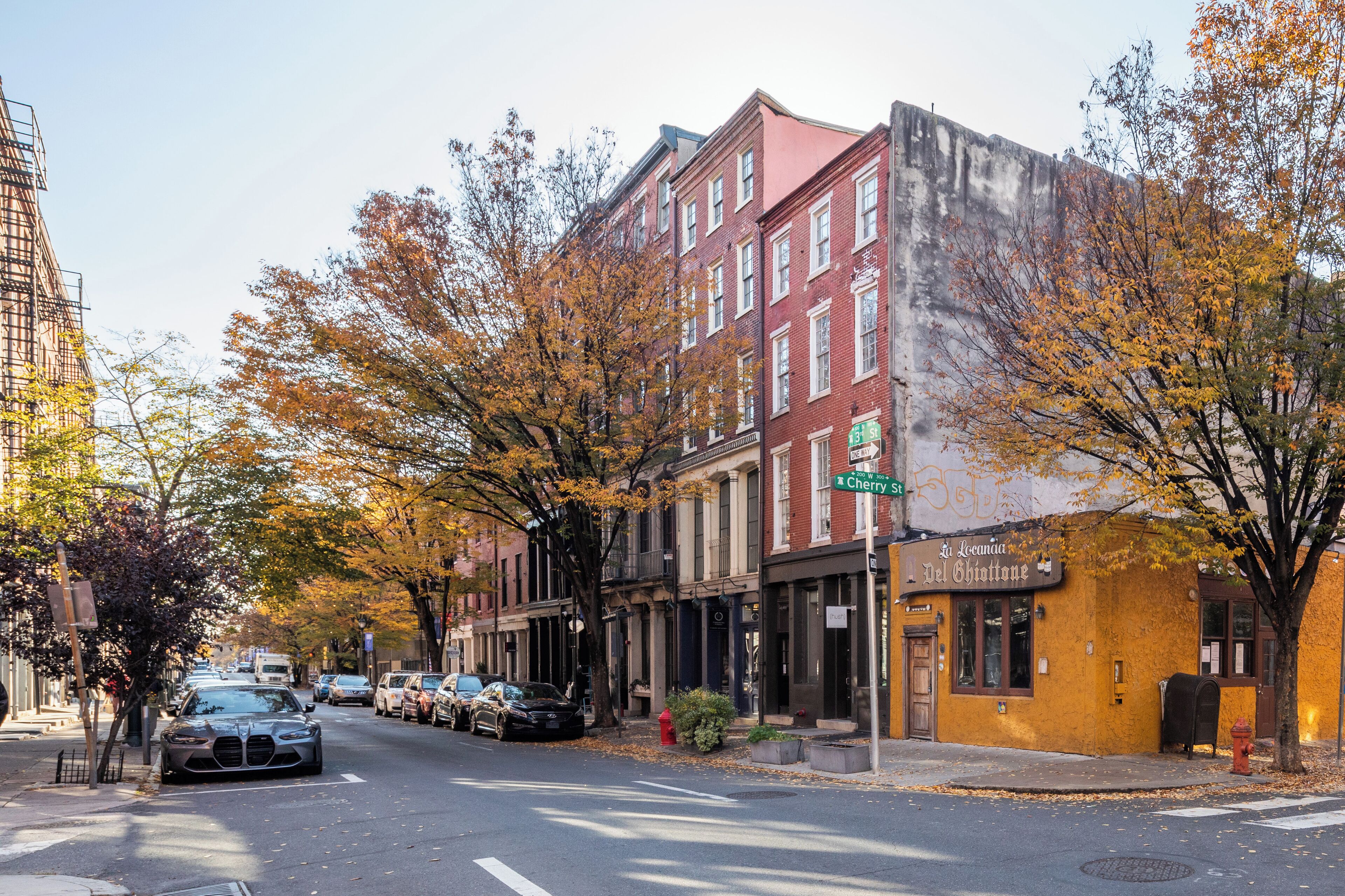 Une vue de rue avec des immeubles, des voitures garées et des arbres d’automne.