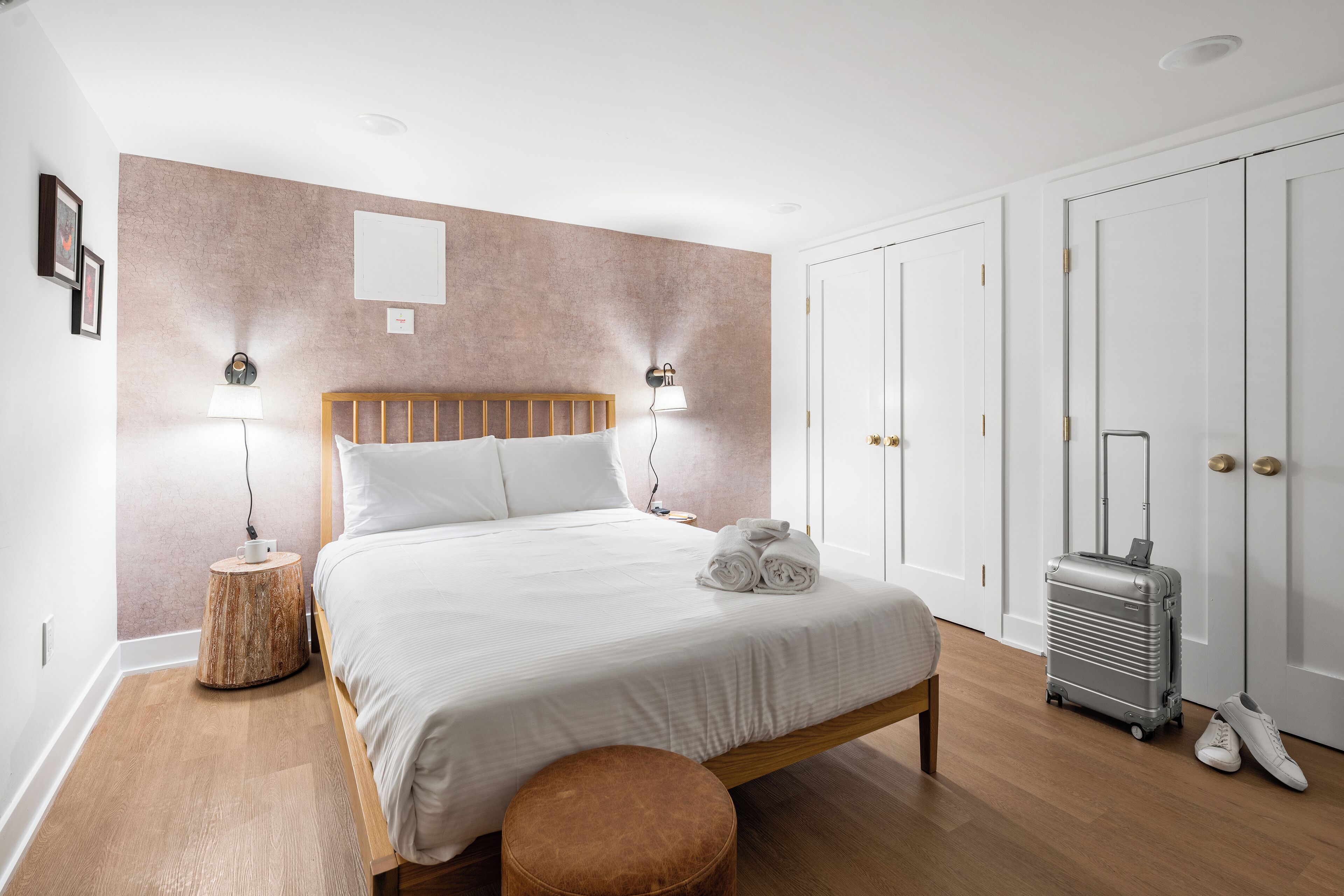 A neatly made bed with white linens, a wooden headboard, and two bedside lamps. A suitcase and a pair of shoes are placed against the wall.