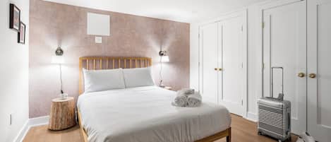 A neatly made bed with white linens, a wooden headboard, and two bedside lamps. A suitcase and a pair of shoes are placed against the wall.