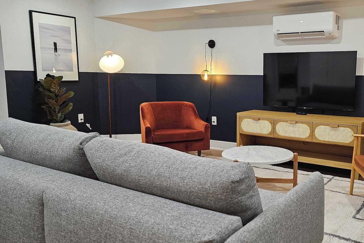 Una sala de estar moderna con un sofá gris, sillas rojas y marrones, una mesa de centro de madera, un televisor de pantalla plana e iluminación empotrada.