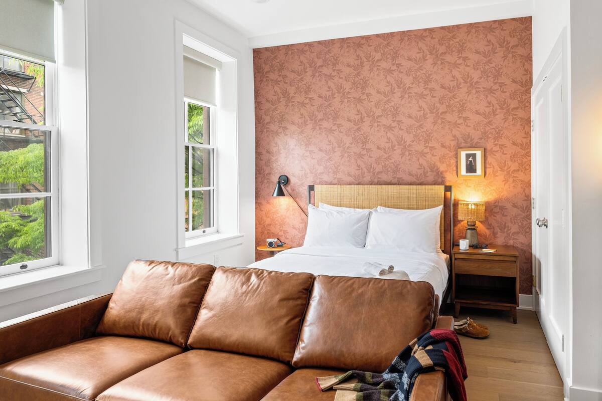 Un dormitorio con cama, sofá de cuero marrón, mesita de noche con lámpara y ventana con vista a follaje.
