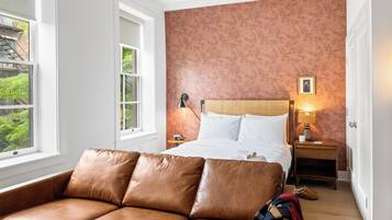 A bedroom with a bed, a brown leather sofa, a nightstand with a lamp, and a window with a view of greenery.