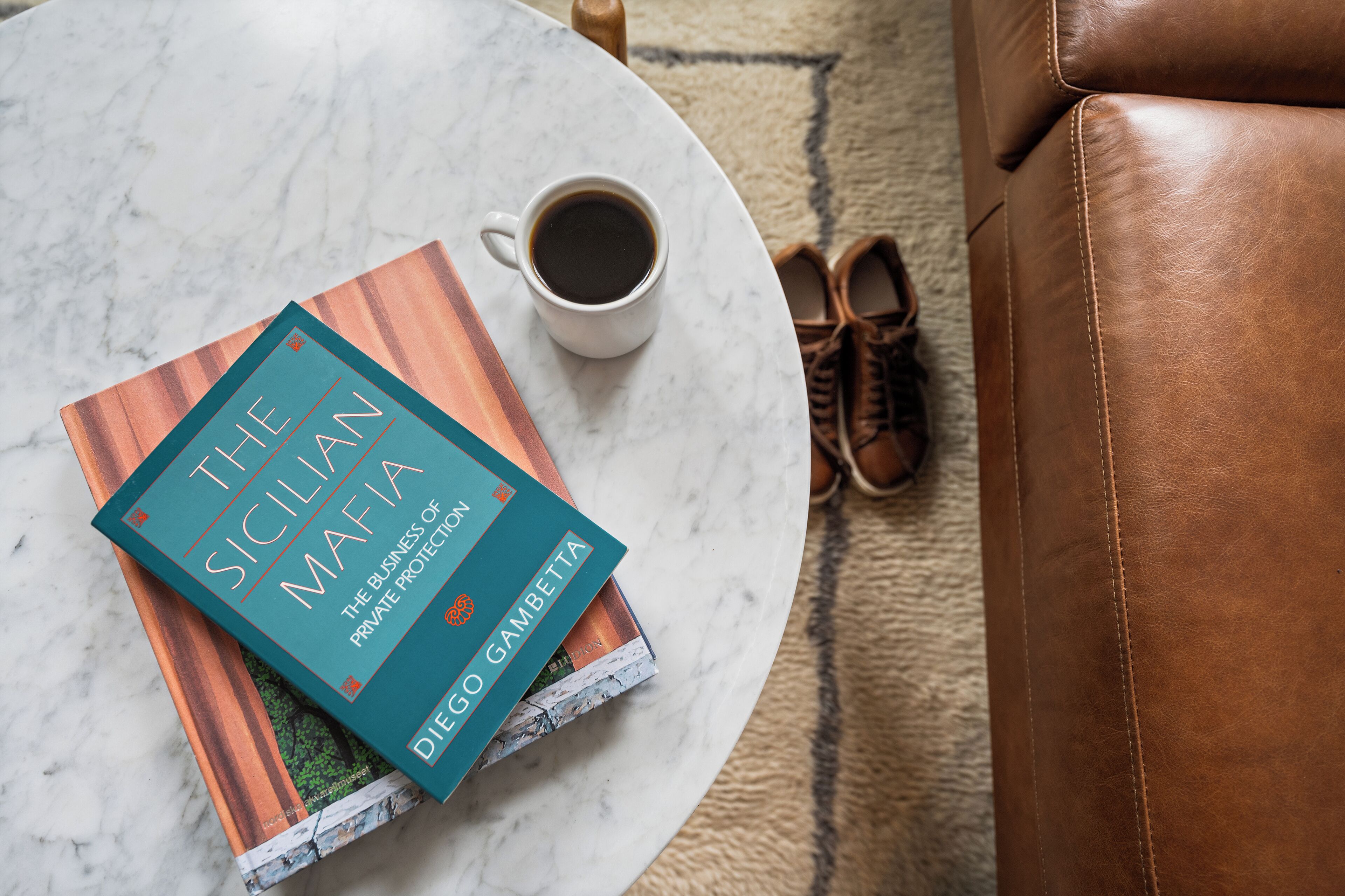 Un libro titulado "La Mafia Siciliana" sobre una mesa redonda, con una taza de café y un par de zapatos al lado.