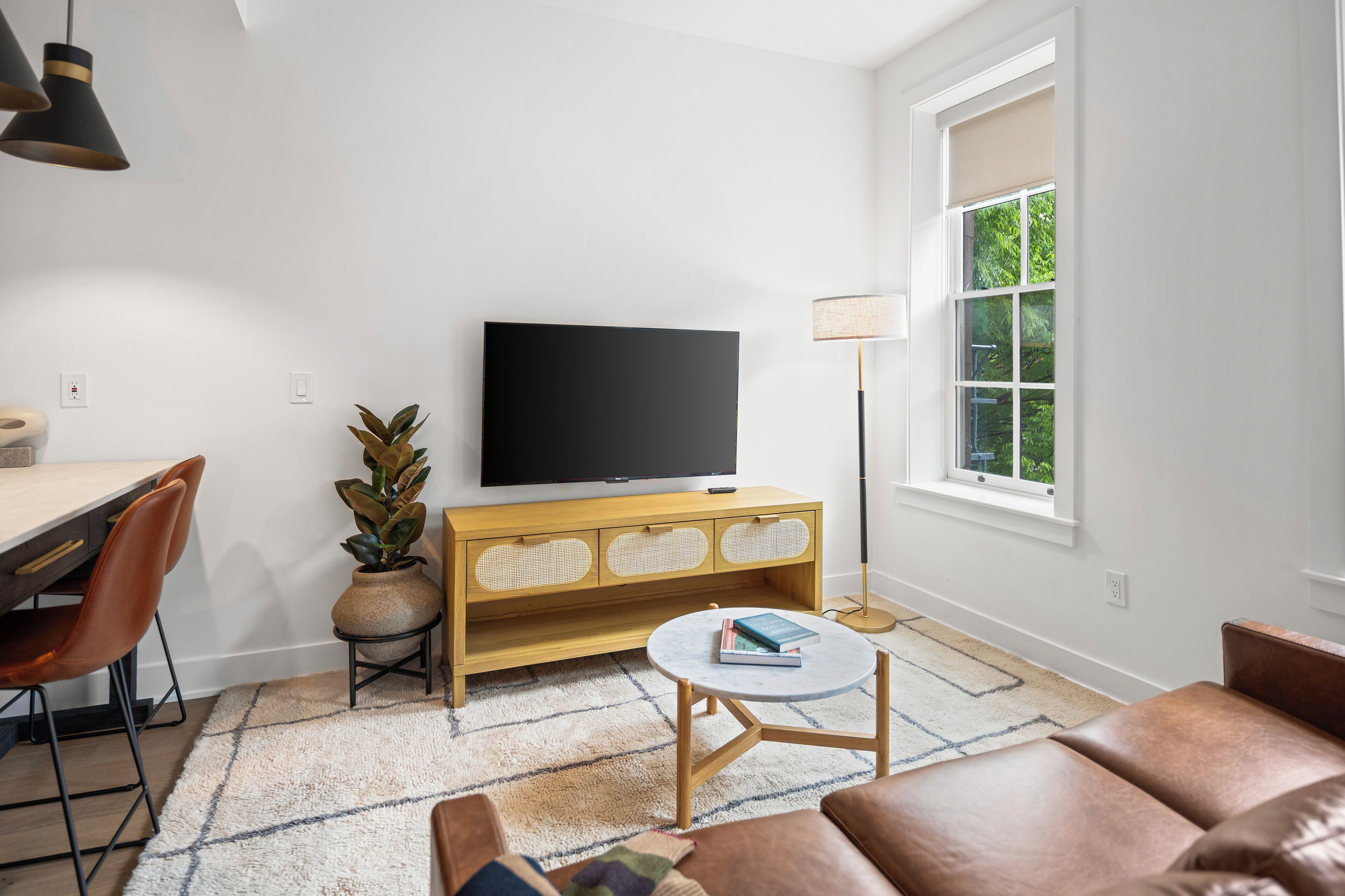 A modern living room with a flat-screen TV, wooden console, leather sofa, and a dining area with chairs and a table.