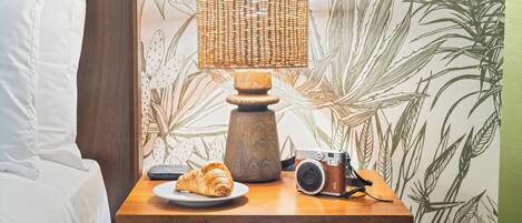A wooden bedside table with a wicker lamp, a camera, and a croissant on a plate.