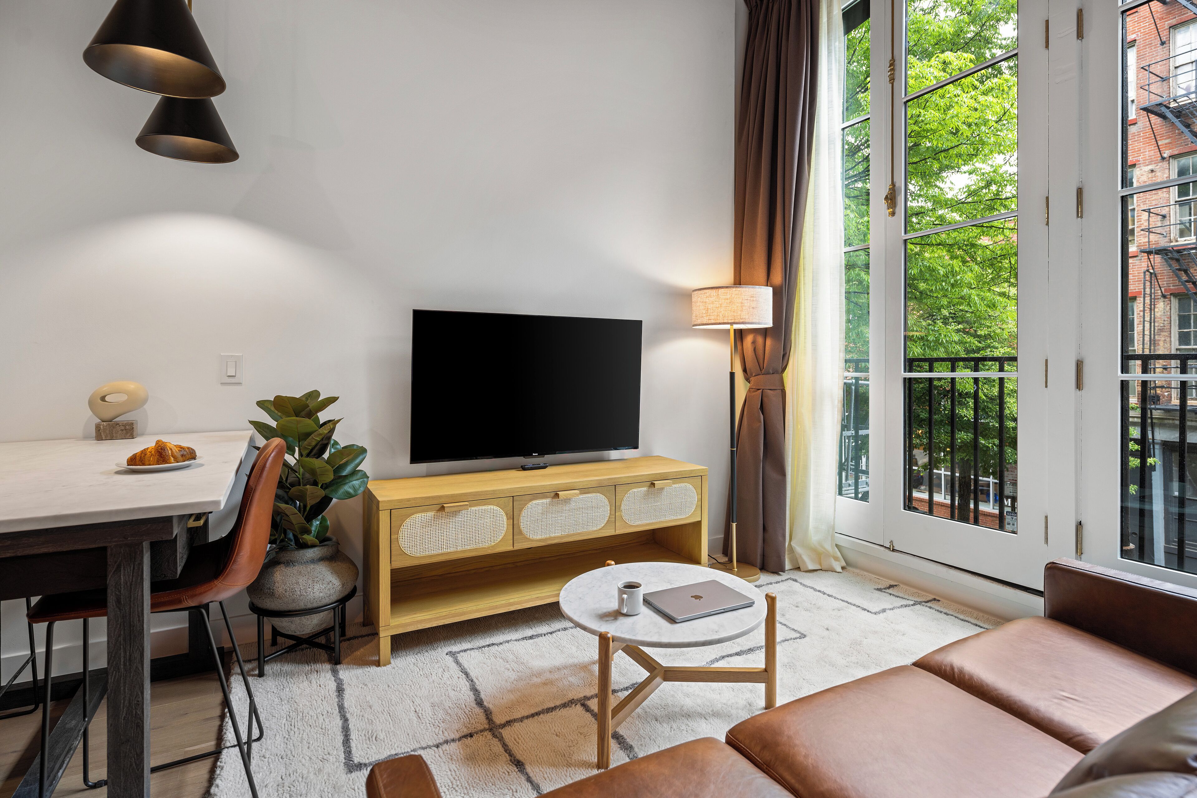 Una sala de estar moderna con una televisión de pantalla plana, una mesa de comedor con sillas, una mesa de centro, un sofá, una planta en maceta y un amplio ventanal con vistas a árboles.