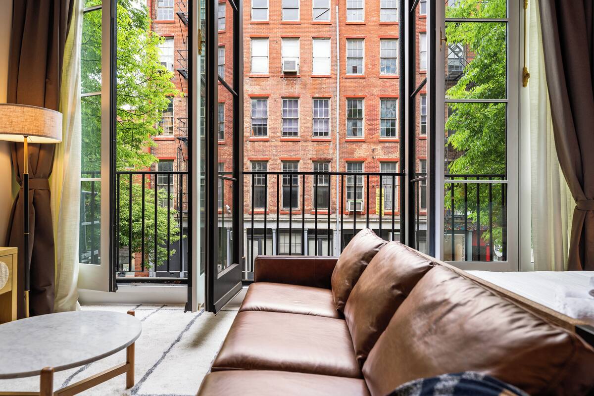 Una sala de estar moderna con un sofá de cuero, una mesa redonda y un amplio ventanal que ofrece vistas a un edificio de ladrillo rojo y árboles verdes.