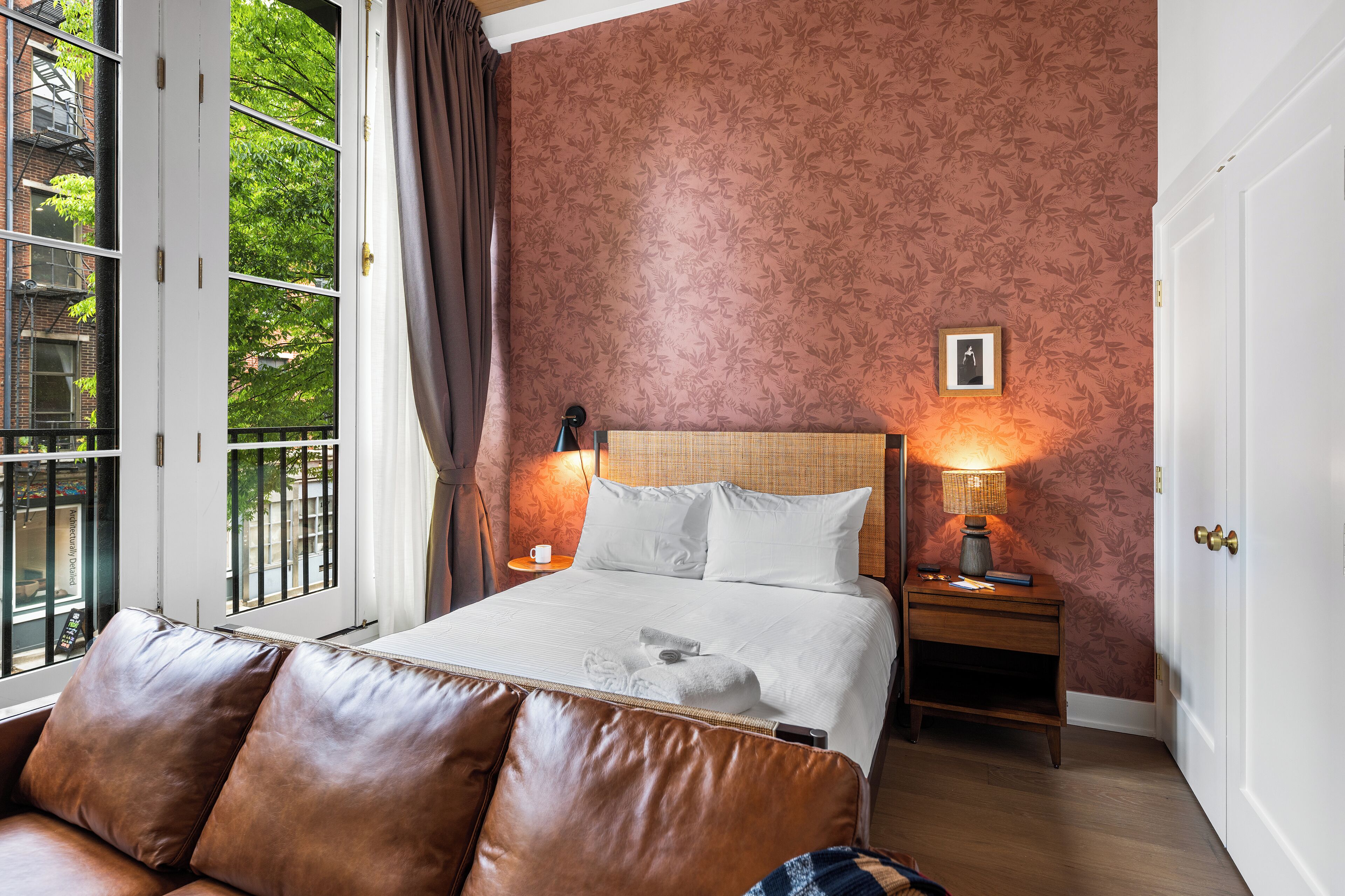 Quarto de hotel com cama, mesa de cabeceira, luminária, janela com cortinas e vista para árvores.