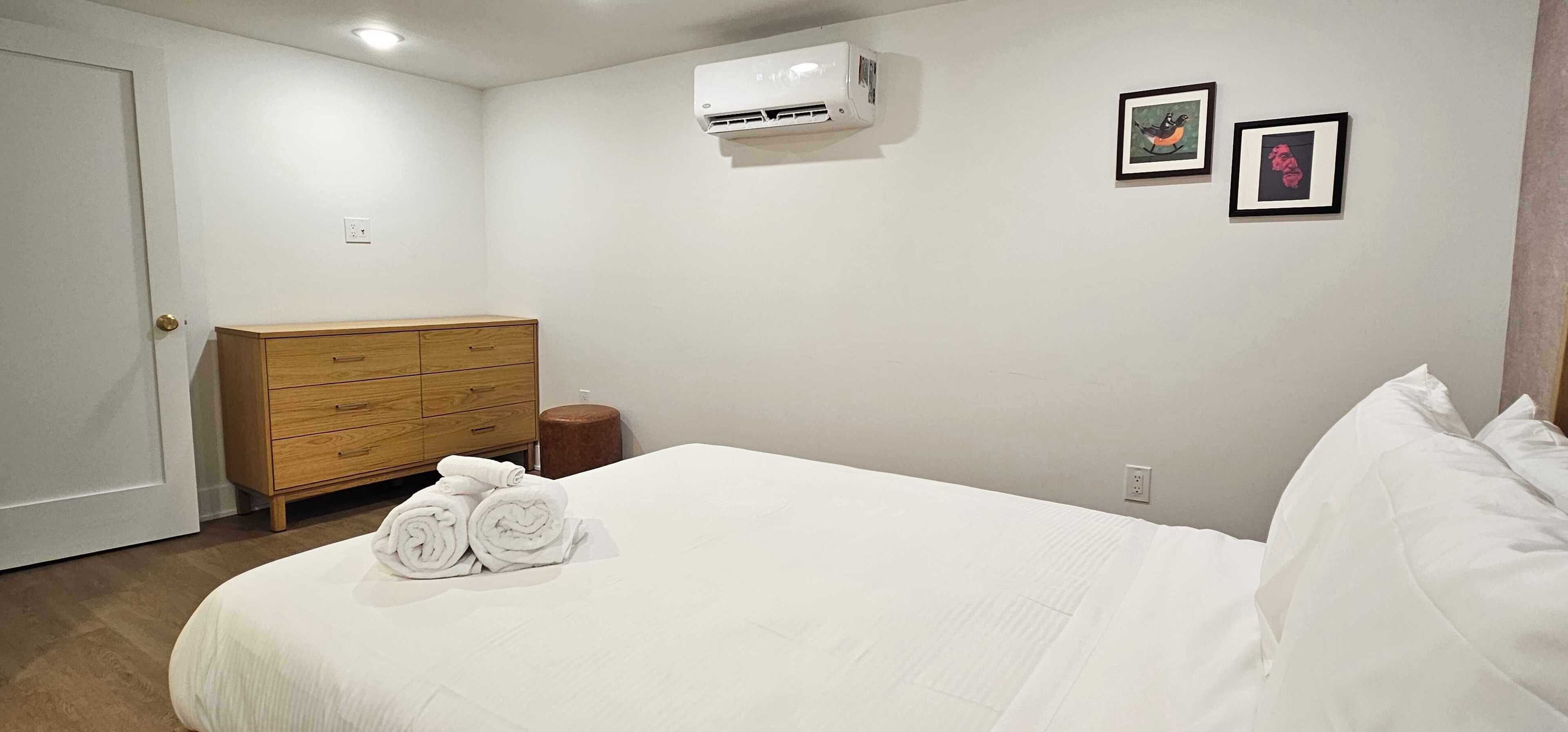 Un dormitorio con cama, cómoda de madera, aire acondicionado y dos cuadros en la pared.