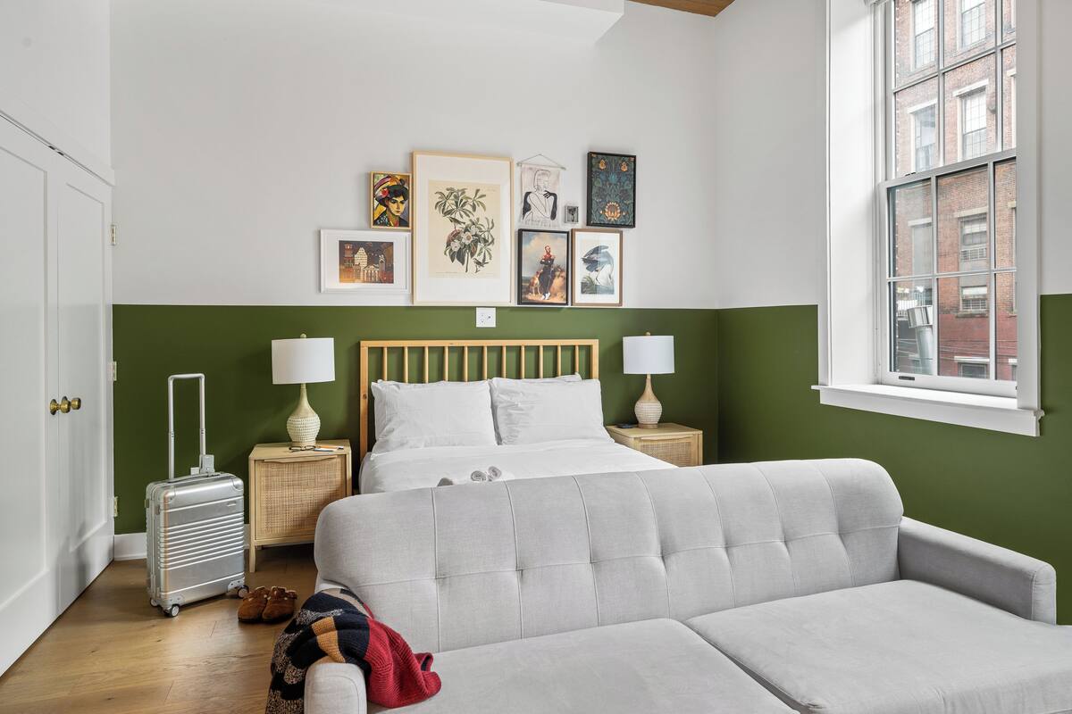 Un dormitorio con cama, mesitas de noche, maleta, sofá y una obra de arte en la pared.