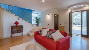 Un salon spacieux avec un canapé rouge, un escalier doté de rampes bleues, une plante en pot et une vue à travers une porte vitrée.
