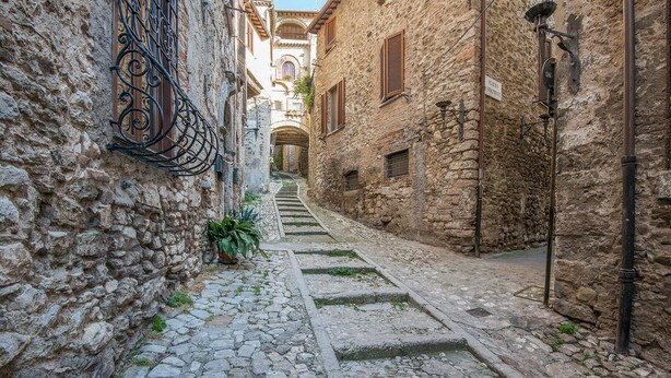 Une ruelle étroite pavée, bordée de bâtiments anciens en pierre, dotés d’arcades et de volets en bois.