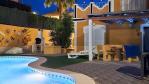 A swimming pool with a curved edge, a patio area with wooden chairs and a table, and a building with a yellow facade and decorative wall art. - Cozy home in Mazarrón (Mazarrón)