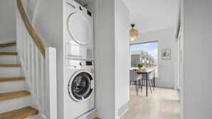 Une buanderie moderne équipée d’un lave-linge et d’un sèche-linge à chargement frontal, d’un escalier en bois et d’un coin repas offrant une vue sur un bâtiment à l’extérieur.