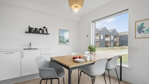 Une cuisine moderne avec des meubles blancs, une table à manger en bois et des chaises grises. On aperçoit un immeuble d’habitation à travers la fenêtre.