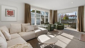 Un salón moderno con un sofá beige, una mesa de centro blanca y dos sillones verdes. Cuenta con un ventanal con cortinas, una obra de arte enmarcada en la pared y vistas al jardín trasero.