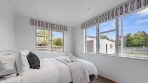 Un dormitorio con cama, almohadas, ventana y vista a una casa.