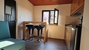 A compact living space with a sofa, a dining area with two stools, a kitchen with cabinets and a refrigerator, and a window providing natural light. - Casa da Capela (Ponte da barca)