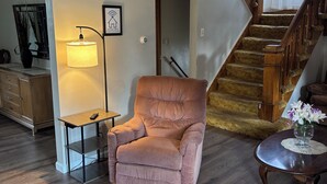 Un salon avec un escalier en bois, un fauteuil confortable, une petite table avec une lampe et un vase rempli de fleurs.