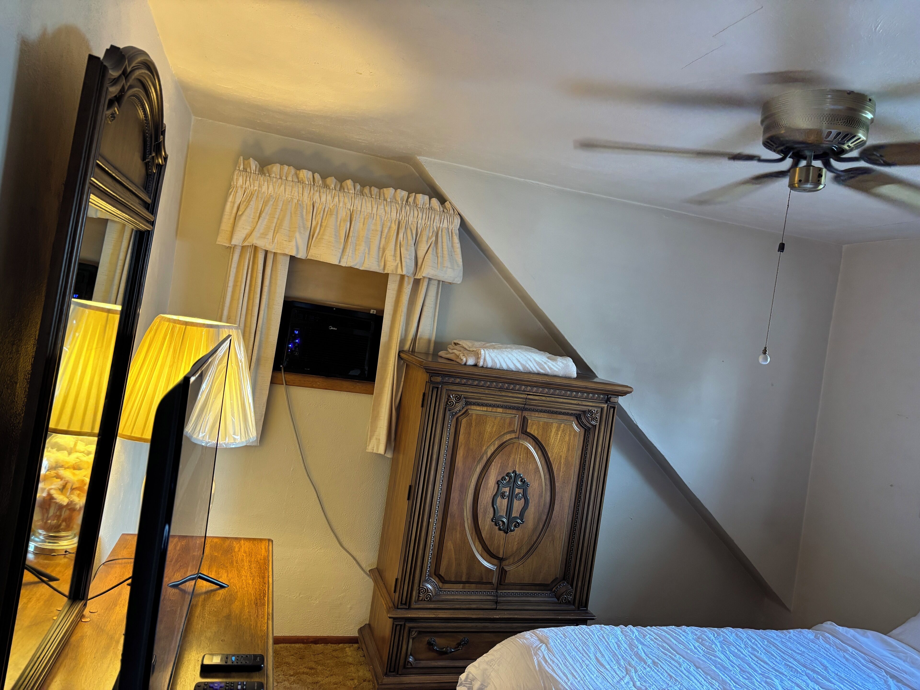 Et soveværelse med skråvæg, ventilator, træskab, seng og vindue med gardiner.
