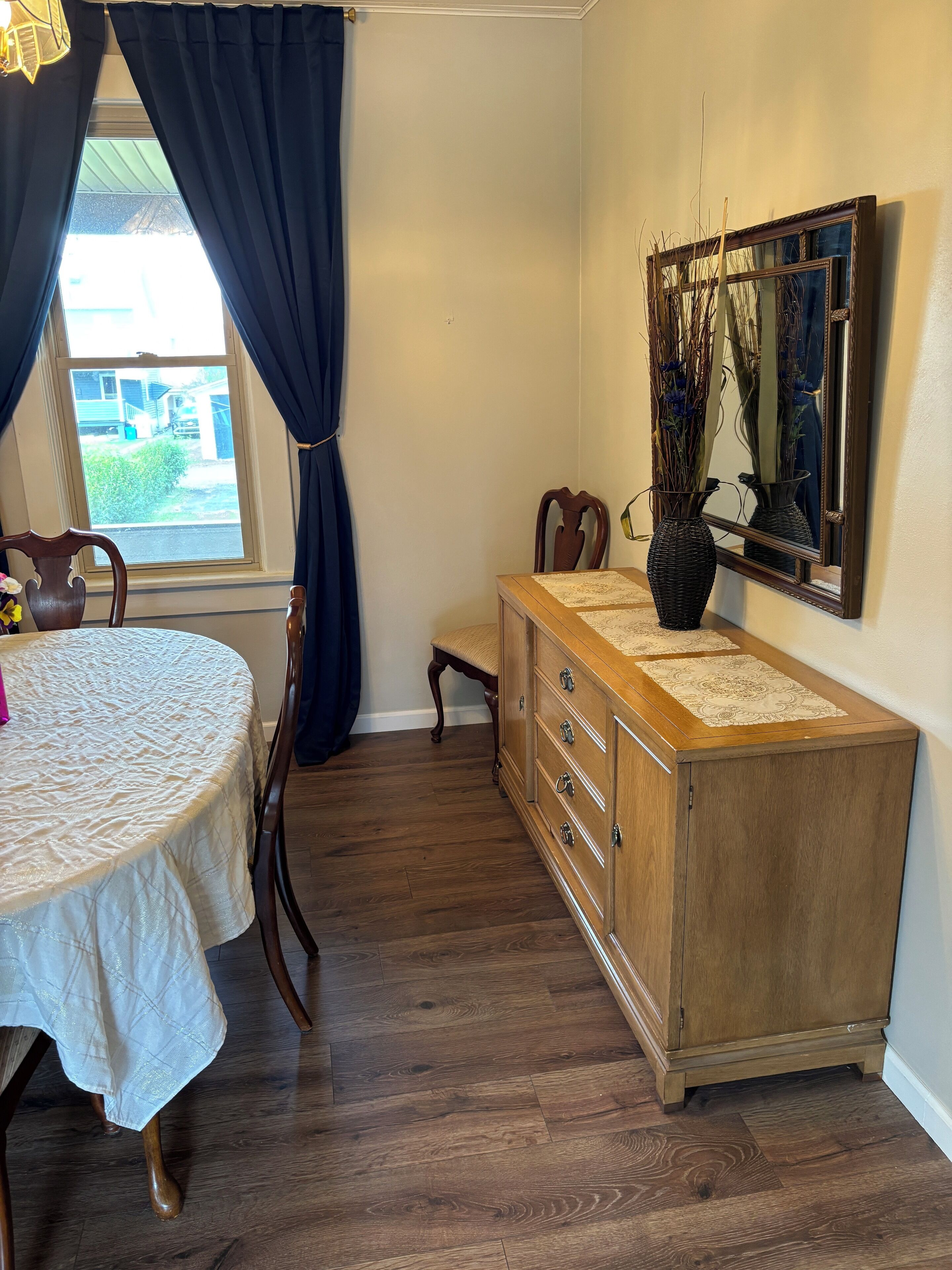 Quarto com cômoda de madeira, mesa de jantar com cadeiras e janela com cortinas.
