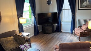 Un salon avec un téléviseur à écran plat, des rideaux foncés, une horloge et un tableau représentant un voilier accroché au mur.