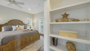 A bedroom with a wooden bed, a ceiling fan, a bookshelf with decorative items, and a window with blinds. - Memory Lane at Magnolia Cottages (Seacrest)