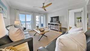 A spacious living room with a ceiling fan, a TV, and a balcony with a view of the beach.