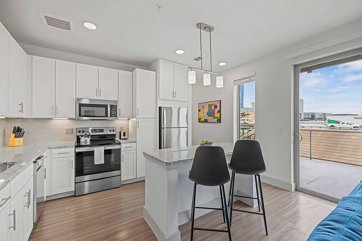 A modern kitchen with white cabinets, stainless steel appliances, and a large island. There are two black bar stools and a view of the city through a large window.